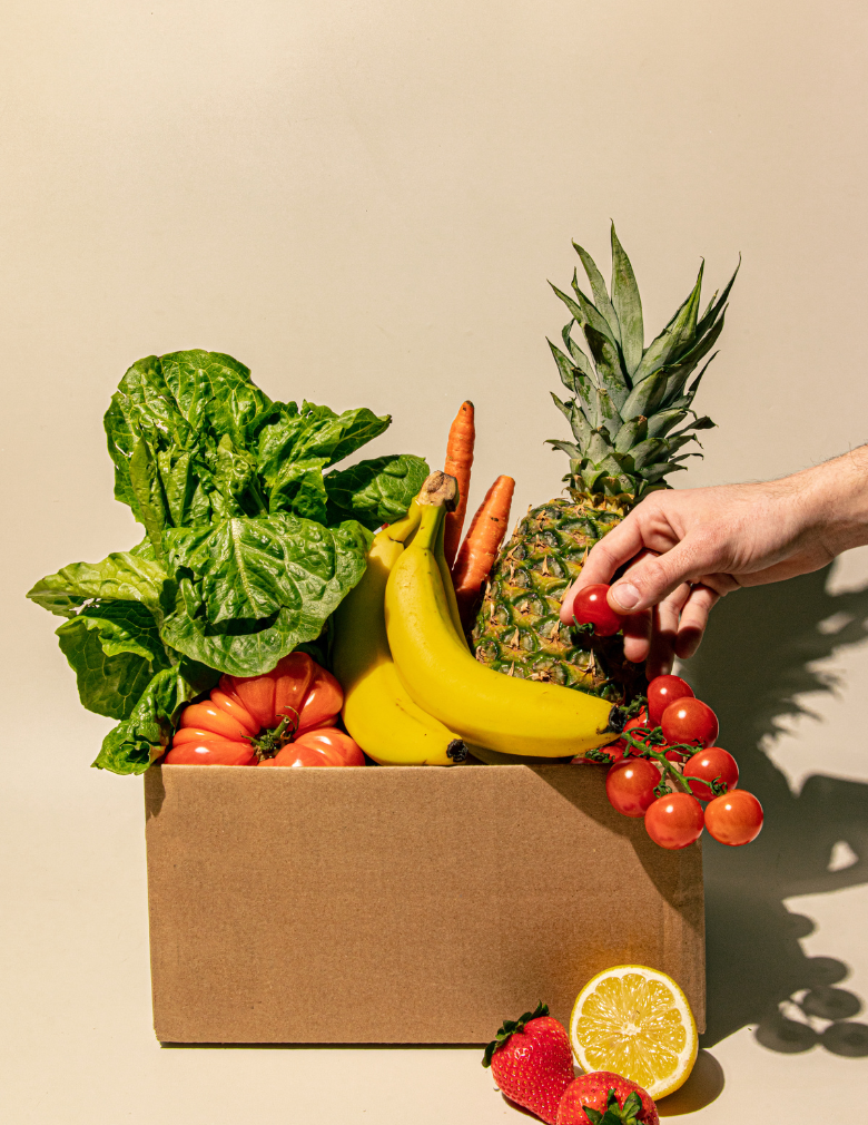Frutas y verdura en una caja de cartón con una mano cogiendo un tomate cherri.