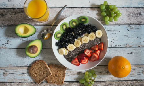 Frutas en un bol sobre una mesa, aguacate sobre la mesa, uvas pan y un zumo de naranja.